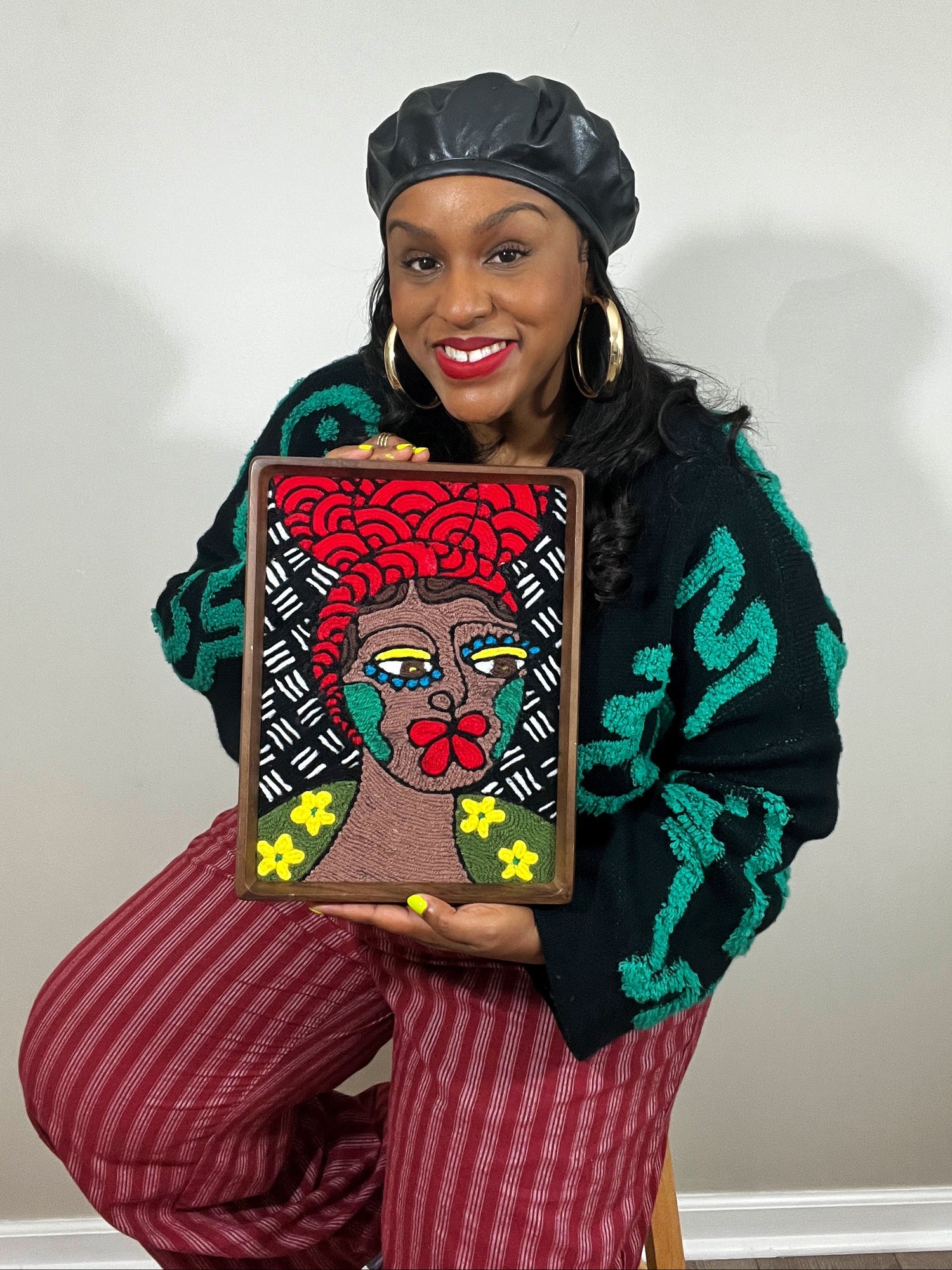 Artist Donna O'Neal holding a framed yarn painting of a stylized female face with bold red lips, yellow eye shadow, blue eye lashes and green organic shapes covering her cheeks; all contrasting beautifully against her medium brown skin. At her shoulders sits a mound of luscious green hills and yellow flowers. Behind her, a black and white mud cloth patterned background.